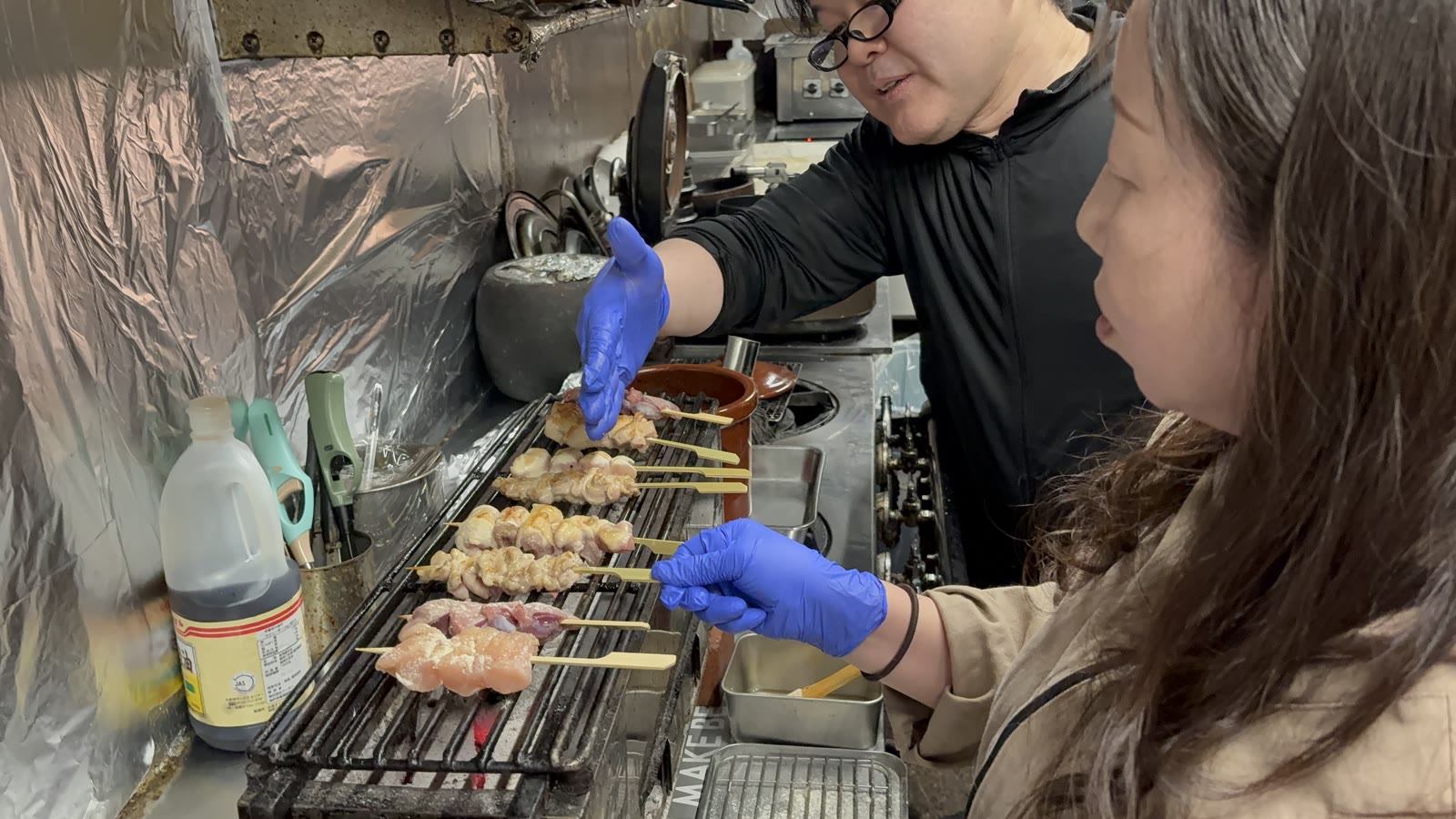 The master guiding a guest grilling yakitori skewers over charcoal at the counter